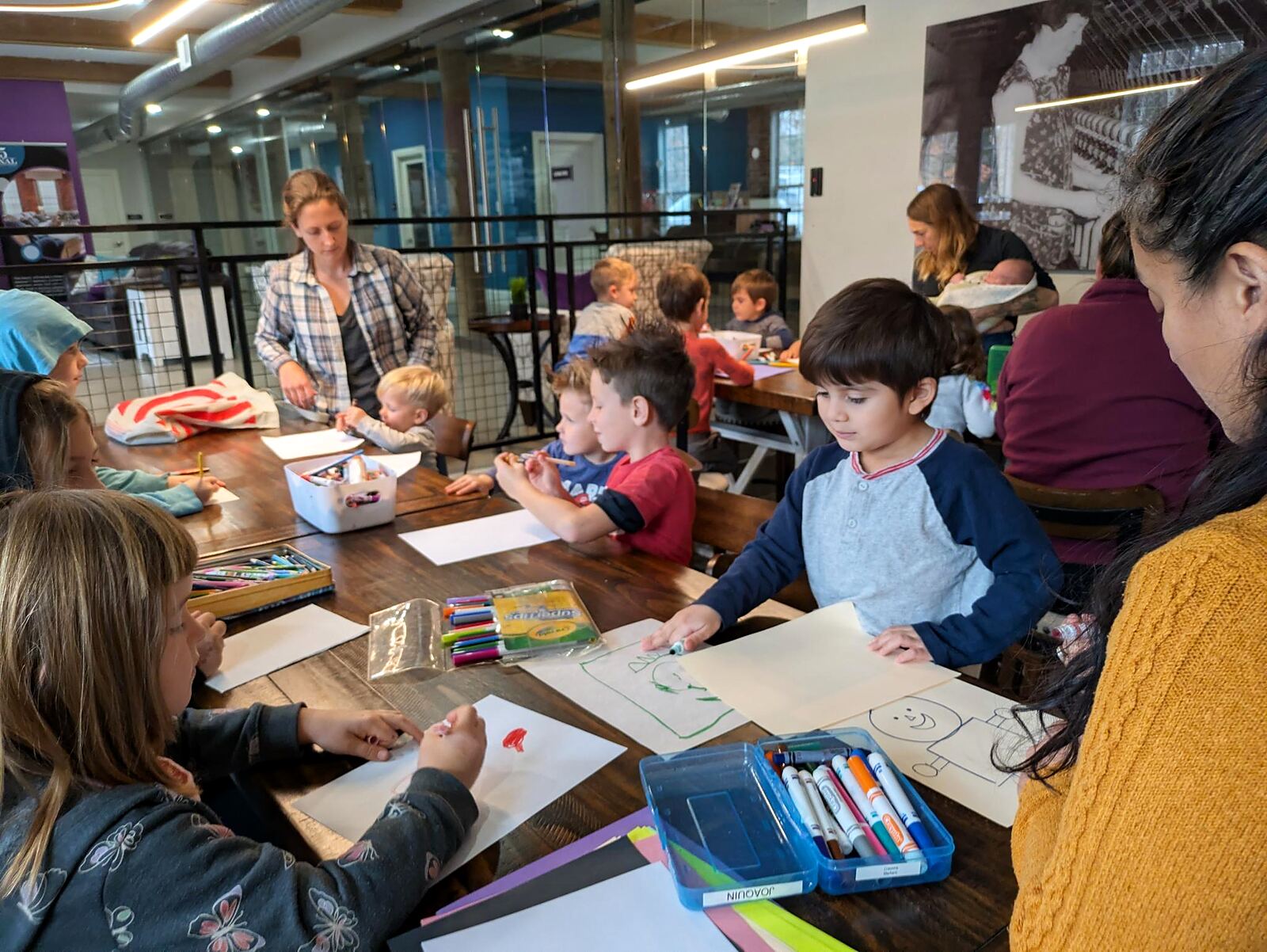 Children at a long table, each drawing and writing on their own sheet, with adults helping.