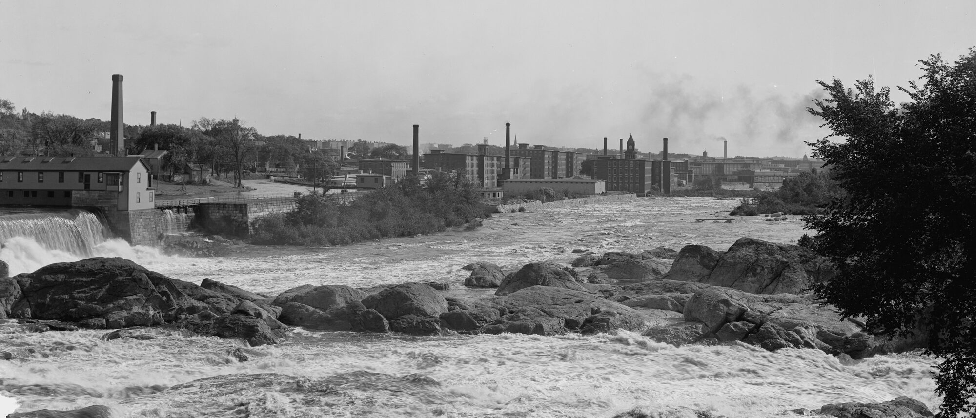 The Merrimack River rushing over rocks below Amoskeag Falls, with the brick mill buildings, smokestacks and clock tower of the Amoskeag mills along the far bank.